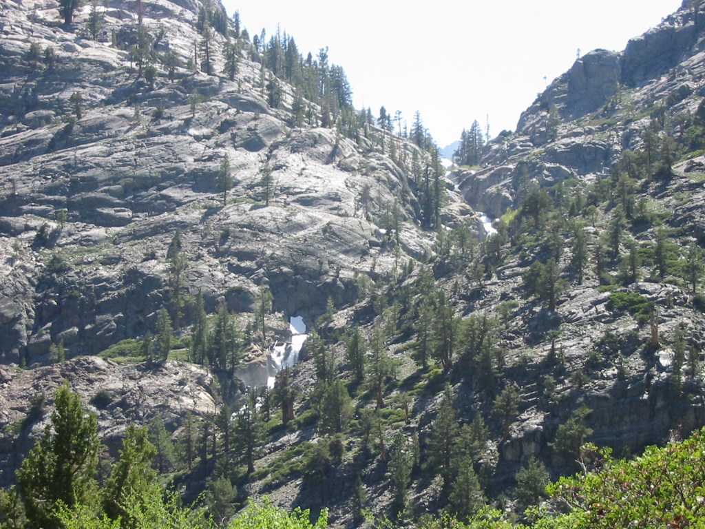 A scenic mountain landscape featuring rocky terrain and scattered pine trees, with a small waterfall flowing down the slope.